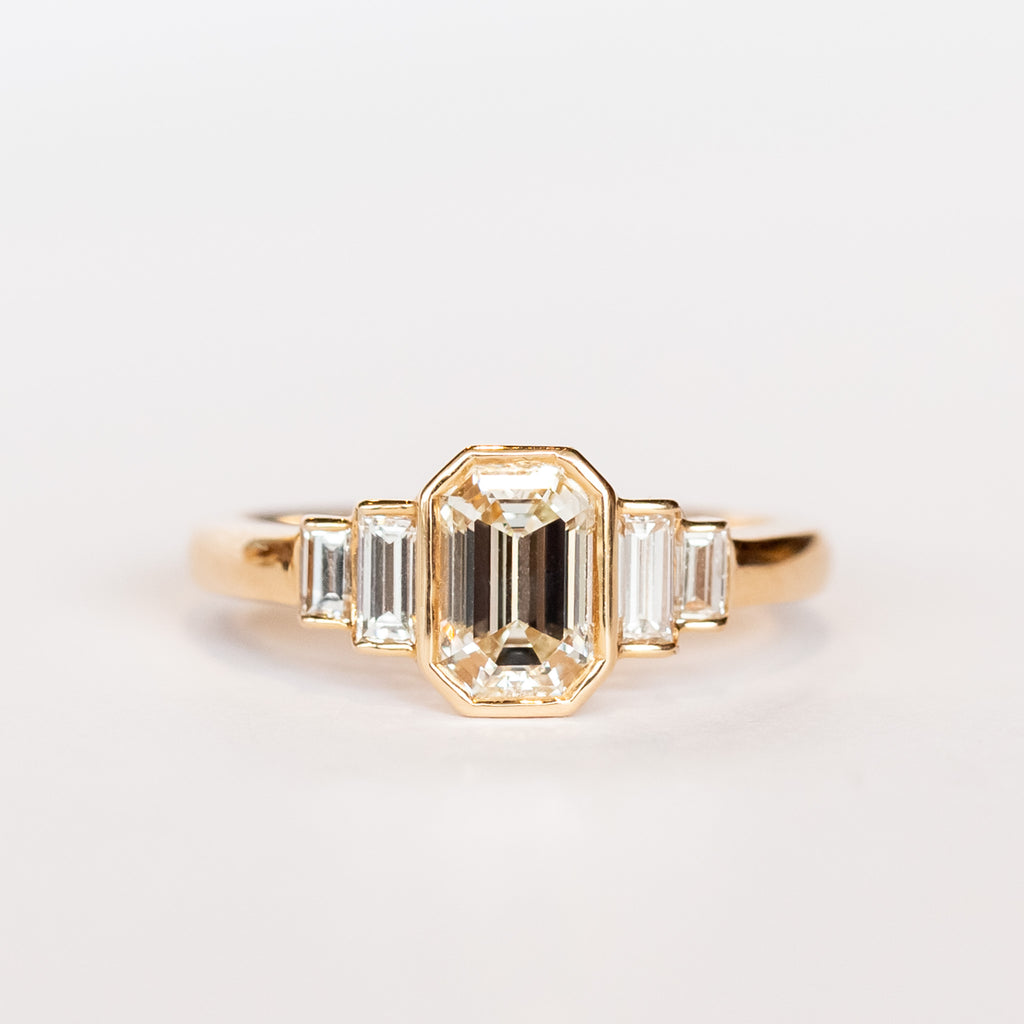 A rose gold engagement ring featuring a bezel set emerald cut diamond center stone flanked by two vertically set baguette diamonds in graduating sizes on either side.