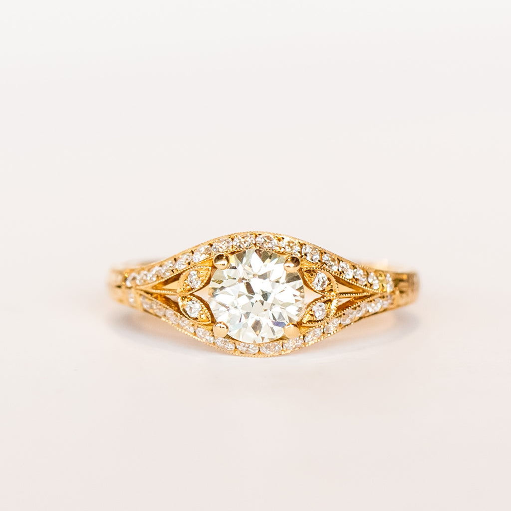 A yellow gold engagement ring that features an old European cut center diamond, floral filigree accents, and rows of pave diamonds.