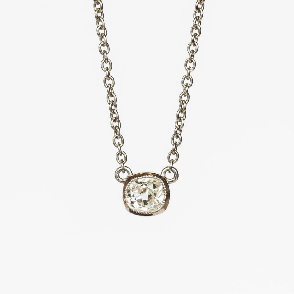 A cushion cut diamond in a platinum bezel setting with milgrain edge, stationed at the center of a platinum cable chain necklace.