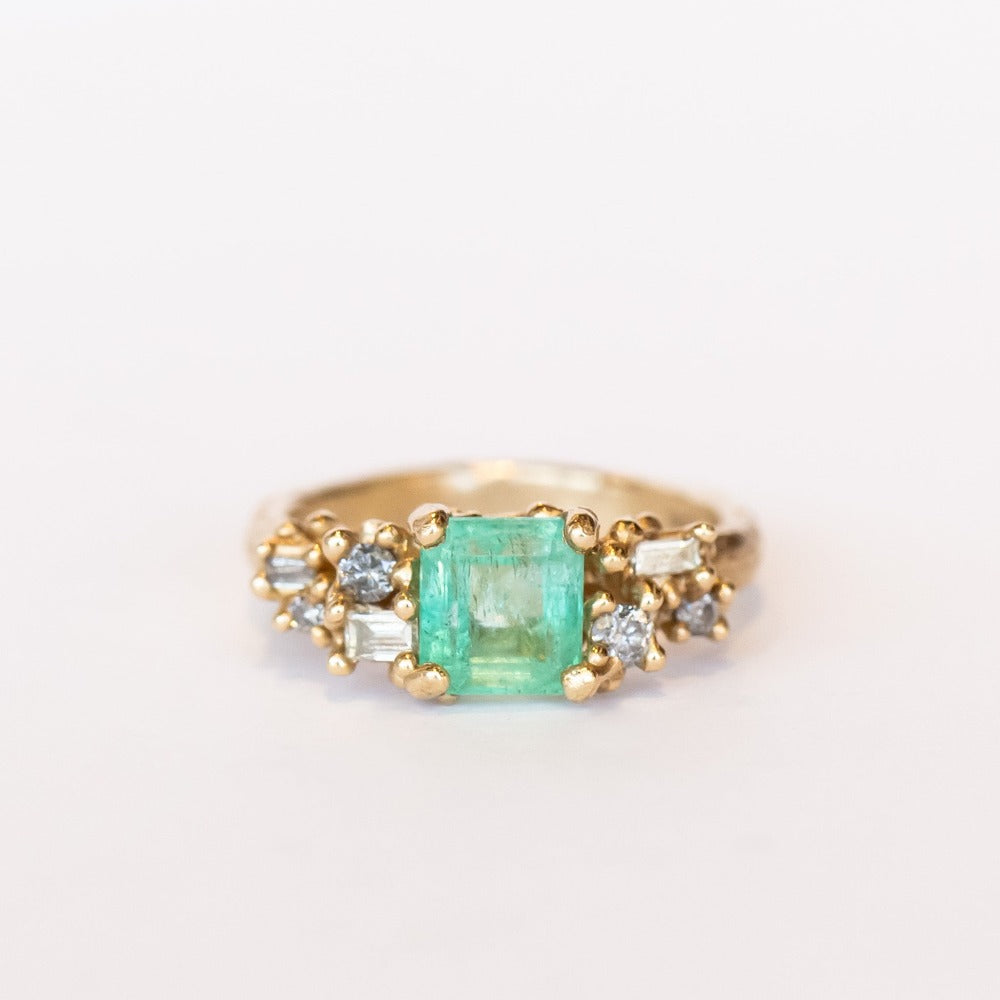 A yellow gold ring featuring an Asscher cut pale green emerald and clusters of diamonds to either side set in granulation.
