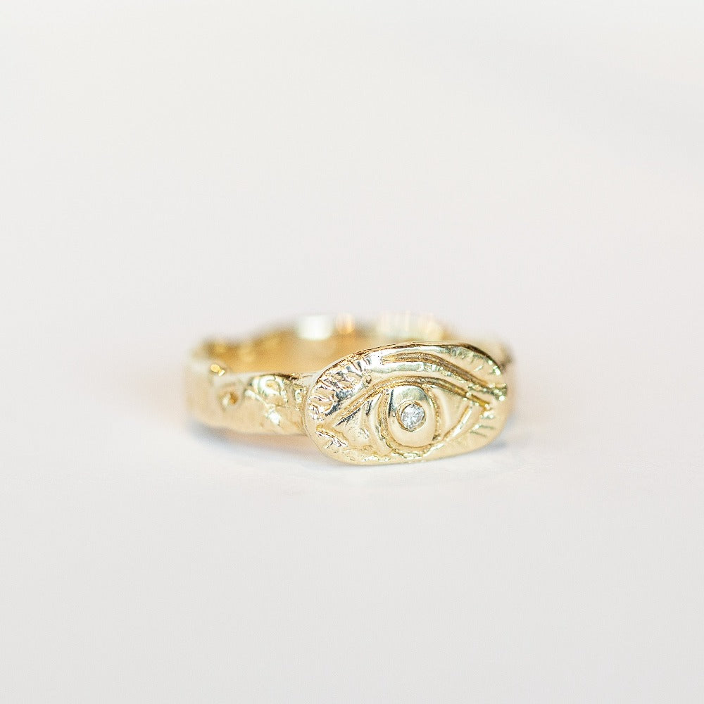A hand-carved yellow gold band, approximately 4mm width, with an oval shaped top plate carved with an eye and a diamond set in its center. slight angle.