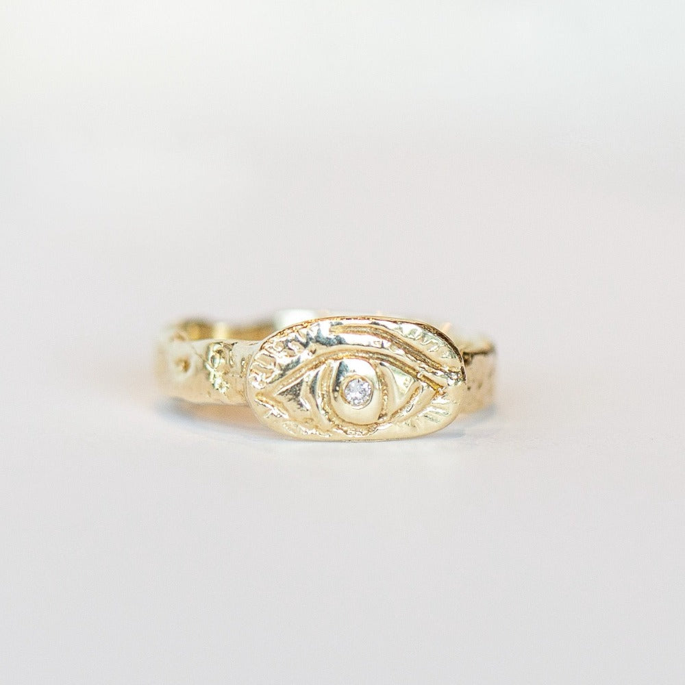 A hand-carved yellow gold band, approximately 4mm width, with an oval shaped top plate carved with an eye and a diamond set in its center. Top view.