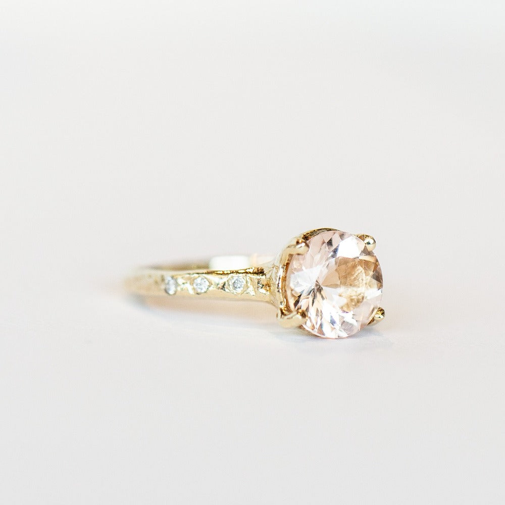 Slight angled view of a hand carved yellow gold Communion by Joy solitaire ring. It features a faceted round morganite gemstone flanked by six small white diamonds on the band.