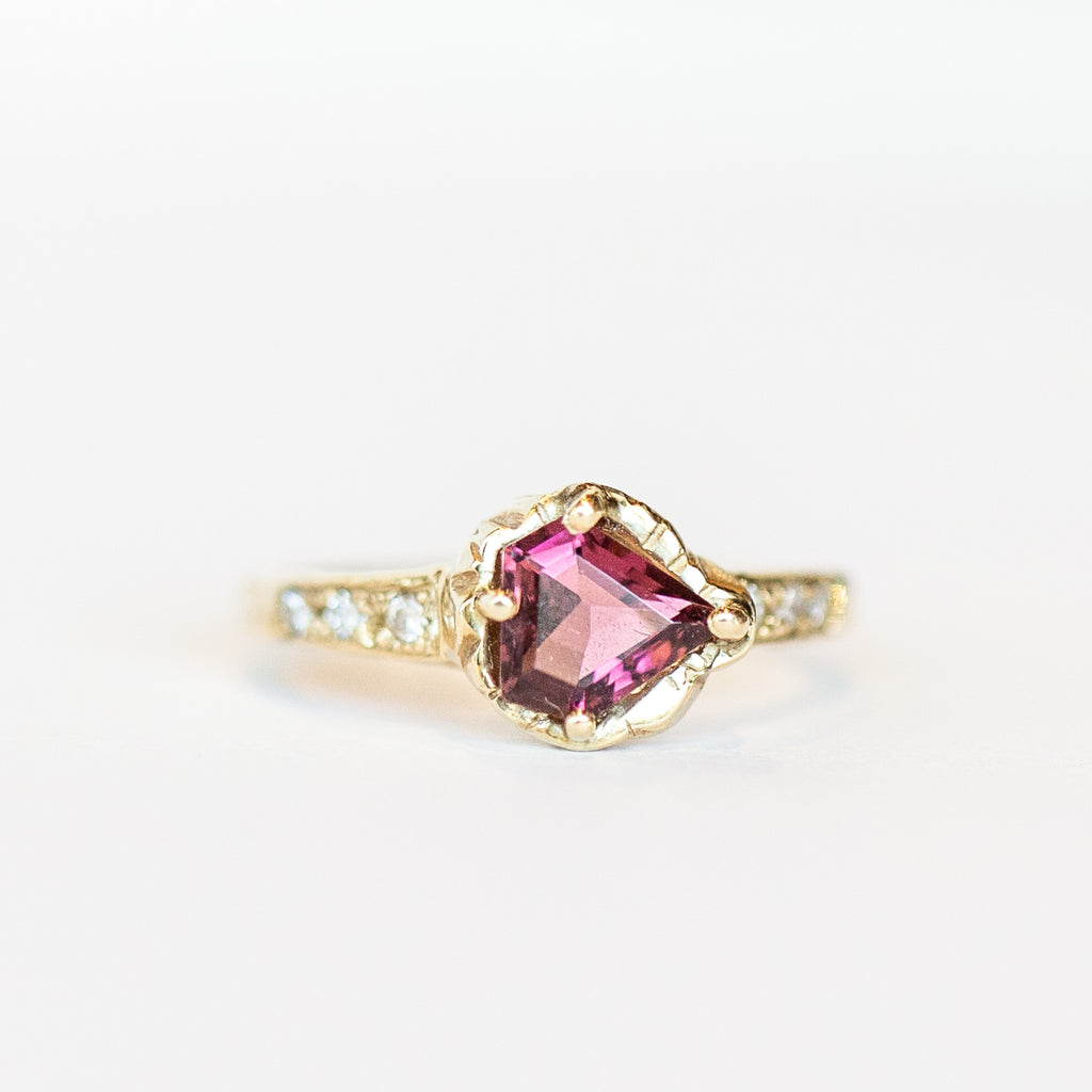 A top view of a hand carved yellow gold Communion by Joy ring featuring a sideways-set shield cut pink tourmaline flanked by six small white diamonds.