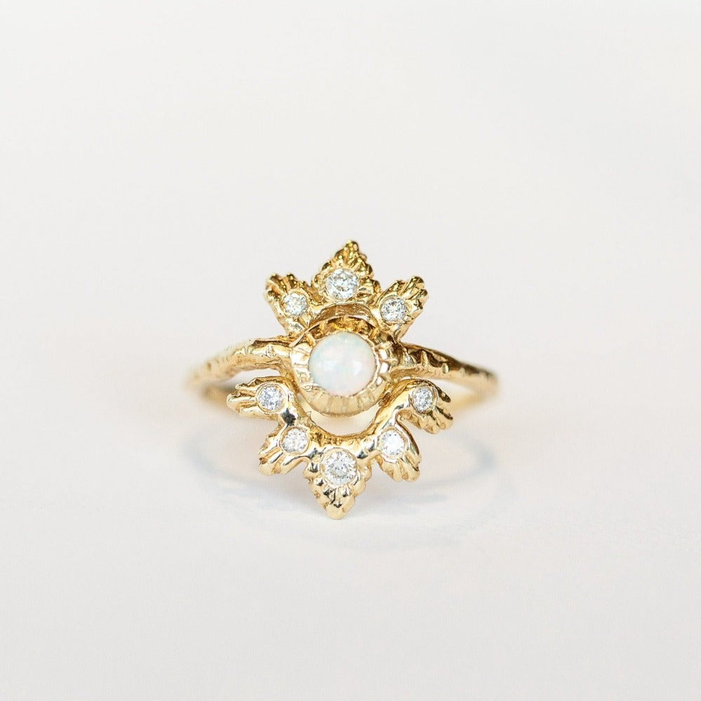 A front view of a yellow gold Communion by Joy ring featuring a delicate band topped with an intricately carved burst shape, centering a bezel set round cabochon opal gemstone and scattered white diamonds.