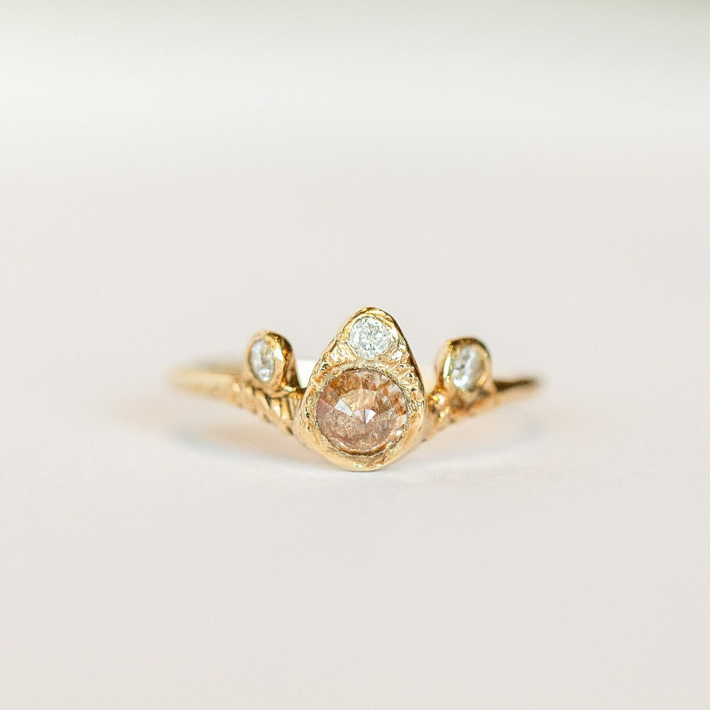 Front view of a Communion by Joy ring that features a rustic, rose cut peach diamond accompanied by three round white brilliant cut diamonds all set in hand carved 14 karat yellow gold.