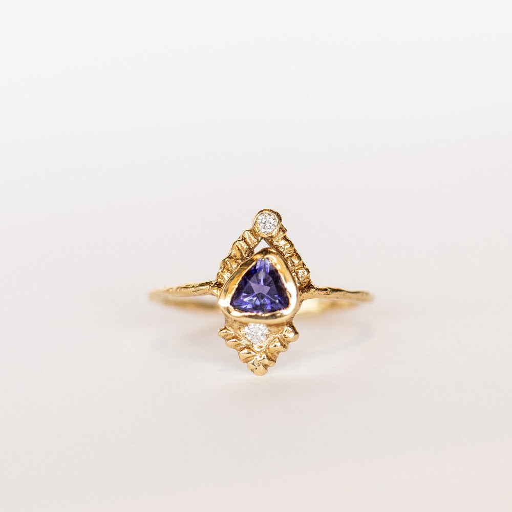 Top view of a yellow gold Communion by Joy ring. It has a thin gold band, with a bluish-purple, trillion cut iolite gemstone bezel set on top, carved pyramid designs and two white diamonds, one at each end.