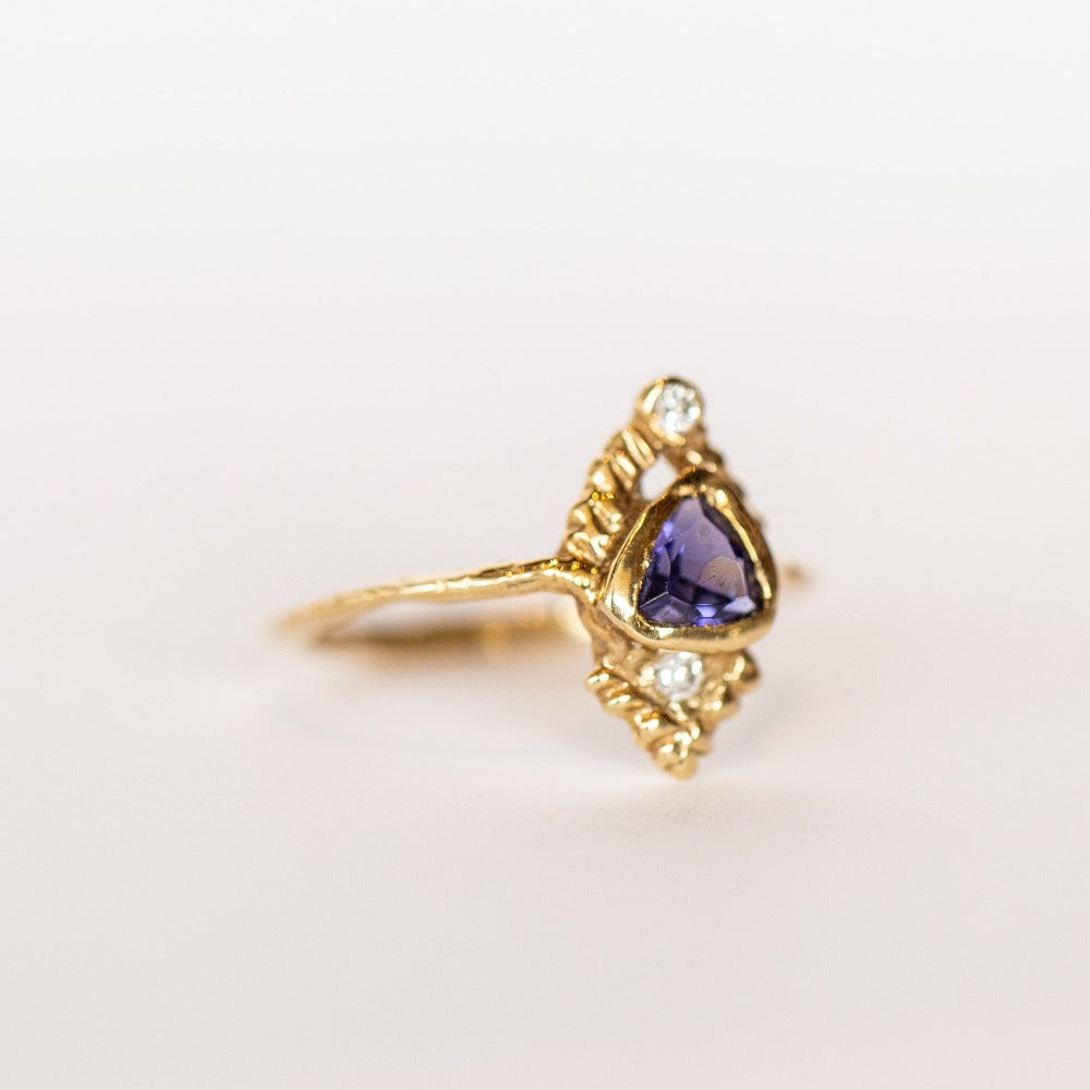 Slight side view of a yellow gold Communion by Joy ring. It has a thin gold band, with a bluish-purple, trillion cut iolite gemstone bezel set on top, carved pyramid designs and two white diamonds, one at each end.