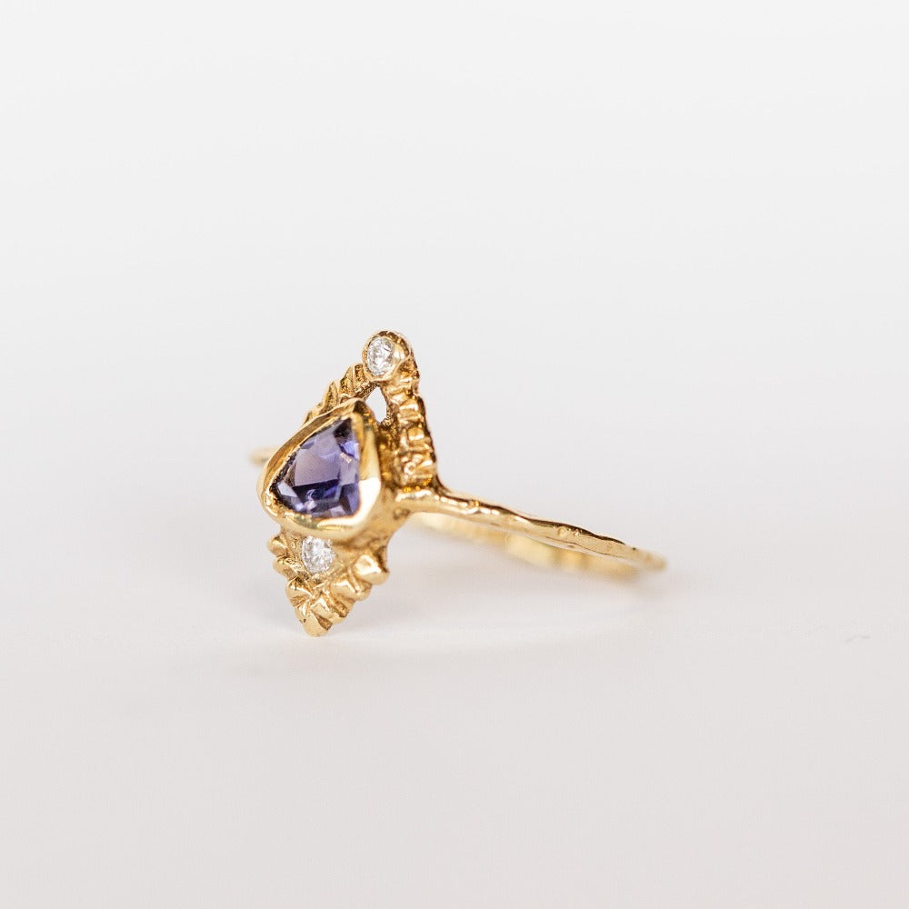 Side view of a yellow gold Communion by Joy ring. It has a thin gold band, with a bluish-purple, trillion cut iolite gemstone bezel set on top, carved pyramid designs and two white diamonds, one at each end.