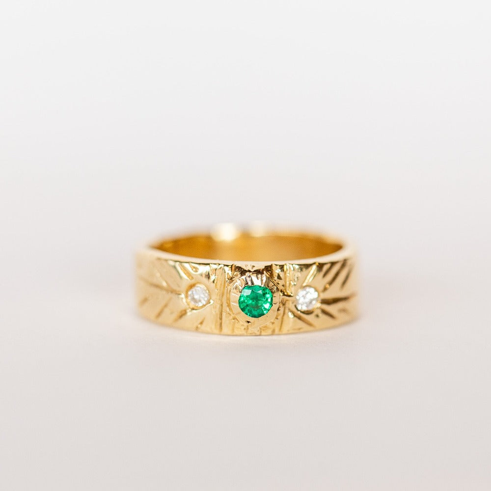 A wide gold band with hand carved detailing set with a center round emerald flanked by two white diamonds.