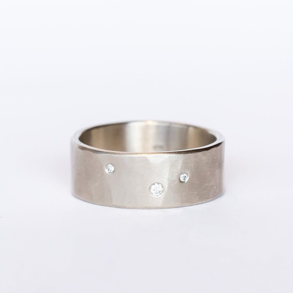 A wide, hammered white gold band with a brushed finish, set with three tiny scattered champagne diamonds.