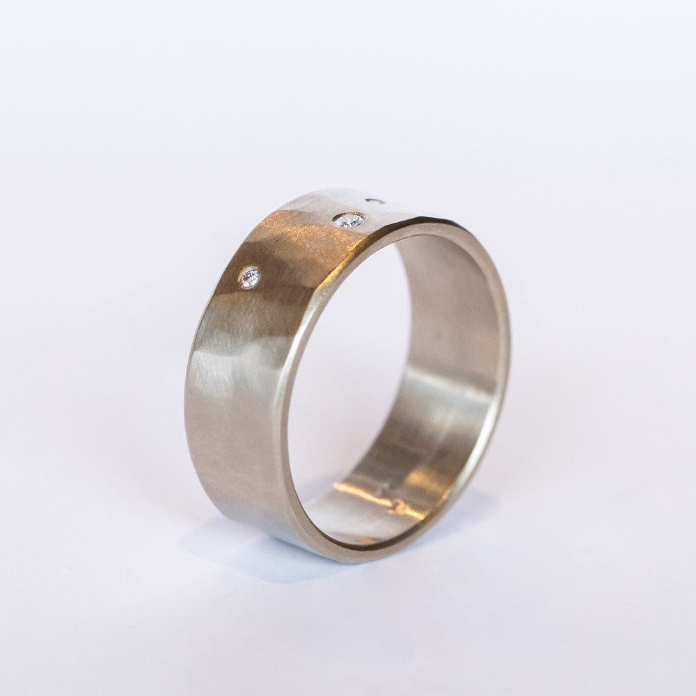 A wide, hammered white gold band with a brushed finish, set with three tiny scattered champagne diamonds.