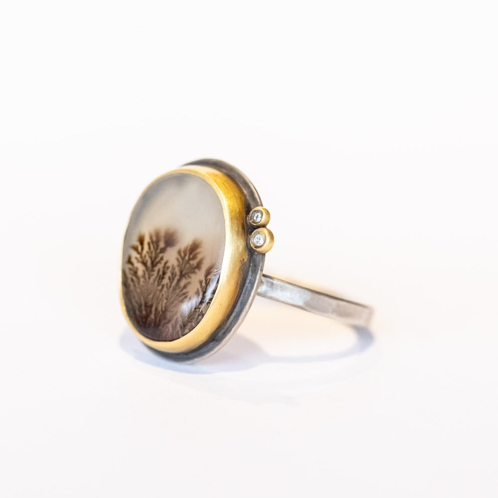 An oval shaped dendritic agate ring with two tiny diamond accents. Set in yellow gold bezel with sterling silver ring. Angle view.