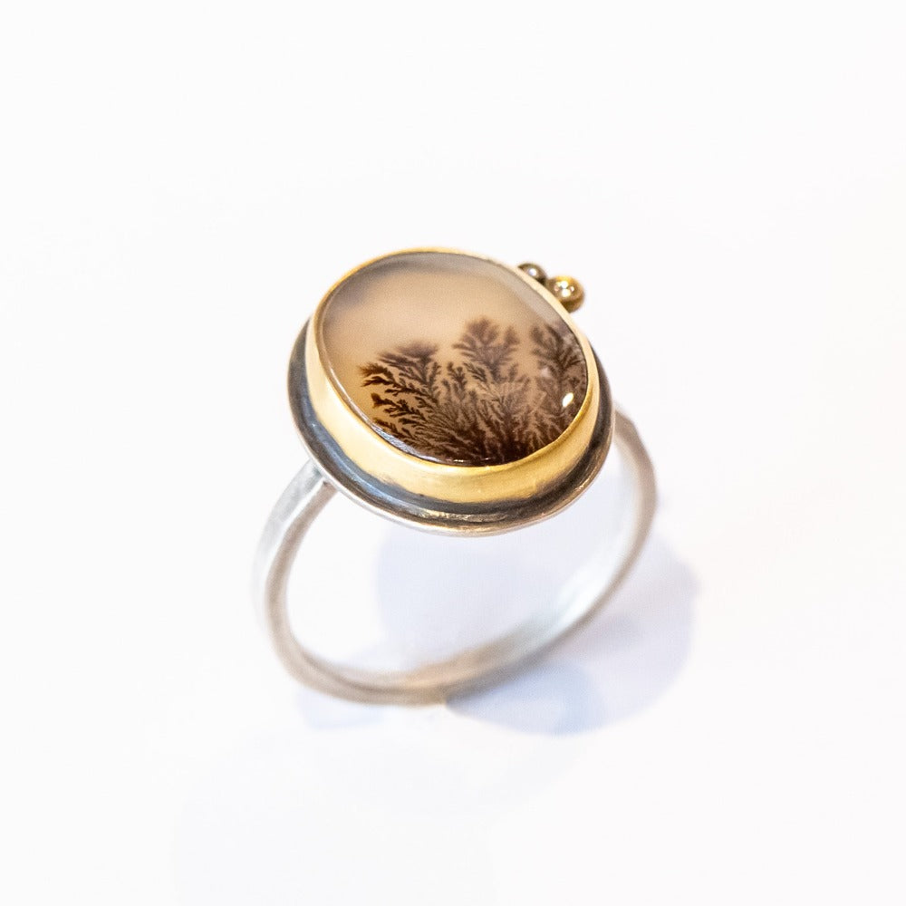 An oval shaped dendritic agate ring with two tiny diamond accents. Set in yellow gold bezel with sterling silver ring. Angle view.