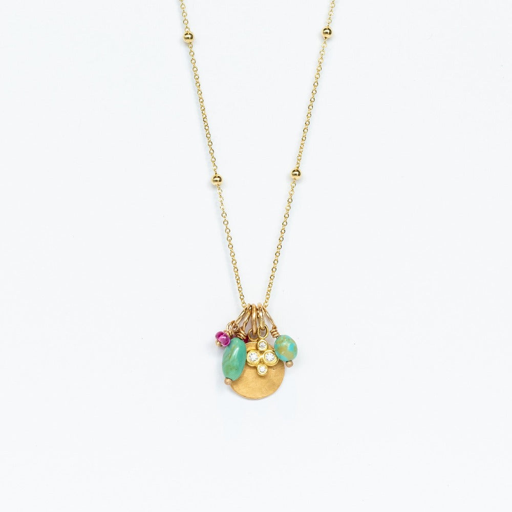 A yellow gold chain necklace with small gold spaced out beads and a handful of charms including a tiny ruby, a hammered gold disc, a diamond clover, and two turquoise beads.
