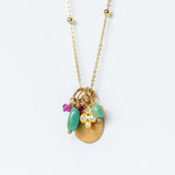 A yellow gold chain necklace with small gold spaced out beads and a handful of charms including a tiny ruby, a hammered gold disc, a diamond clover, and two turquoise beads.