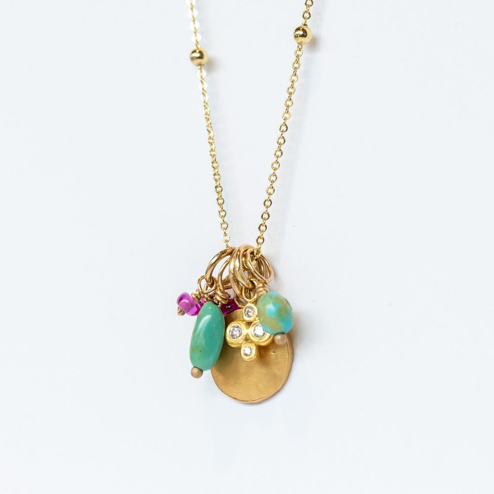 A yellow gold chain necklace with small gold spaced out beads and a handful of charms including a tiny ruby, a hammered gold disc, a diamond clover, and two turquoise beads.