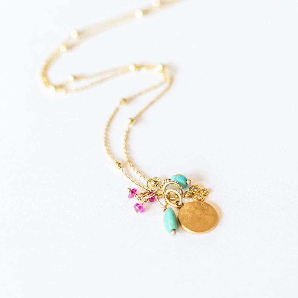 A yellow gold chain necklace with small gold spaced out beads and a handful of charms including a tiny ruby, a hammered gold disc, a diamond clover, and two turquoise beads.