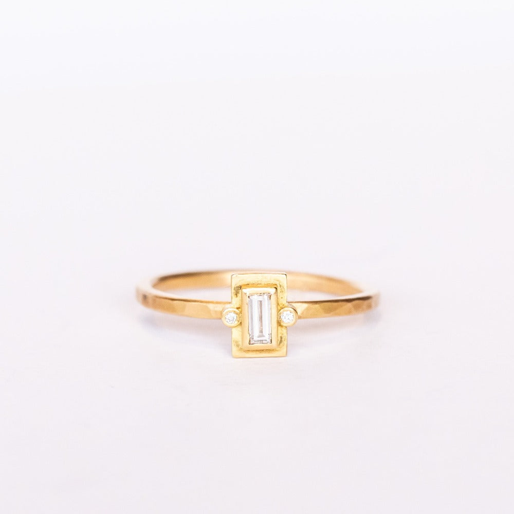 A thin hammered gold ring set with a tiny, bezel-set baguette diamond flanked by two tiny round diamonds.