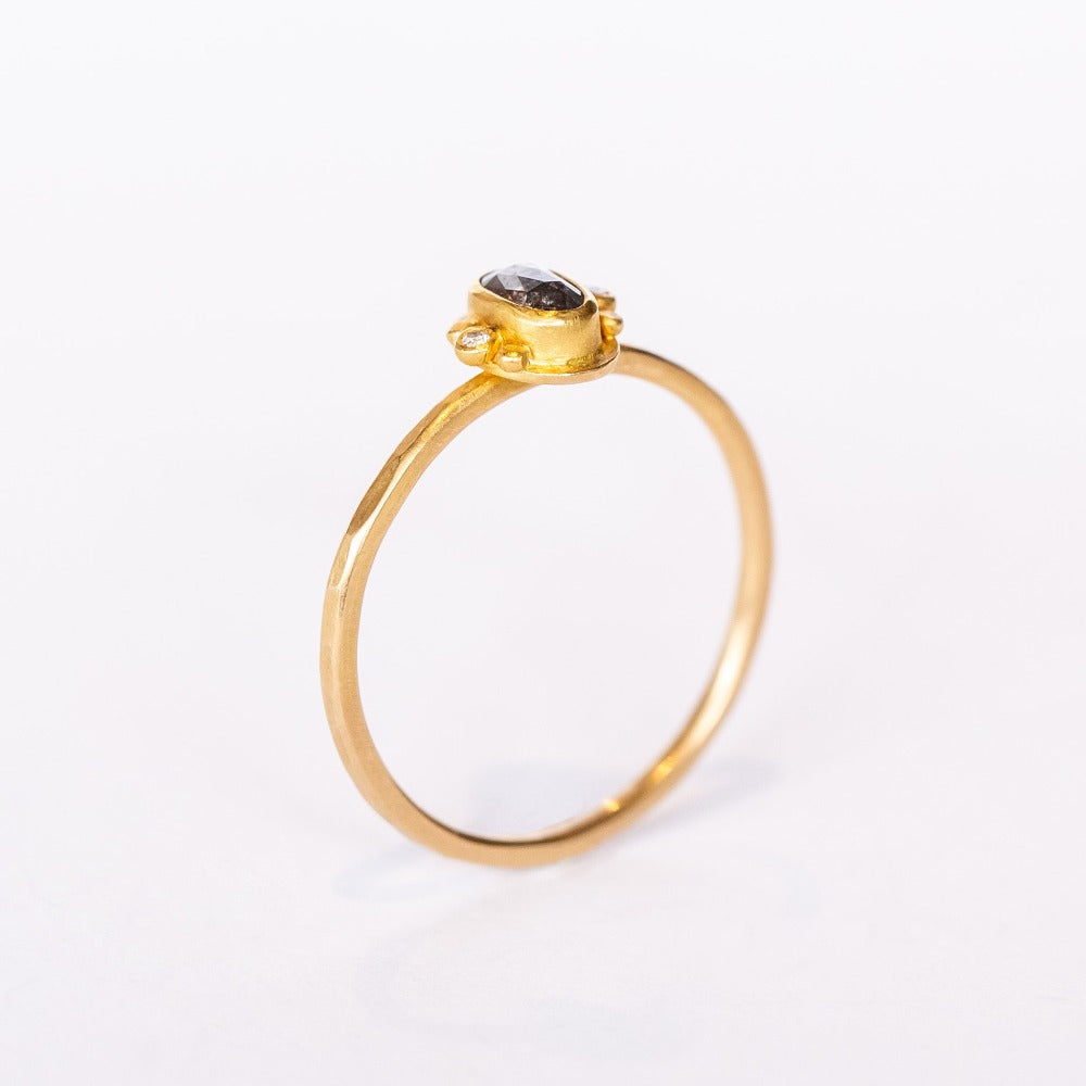 A tiny oval rose cut salt and pepper diamond is bezel set, flanked by two tiny white diamonds and dot details on a thin gold band.