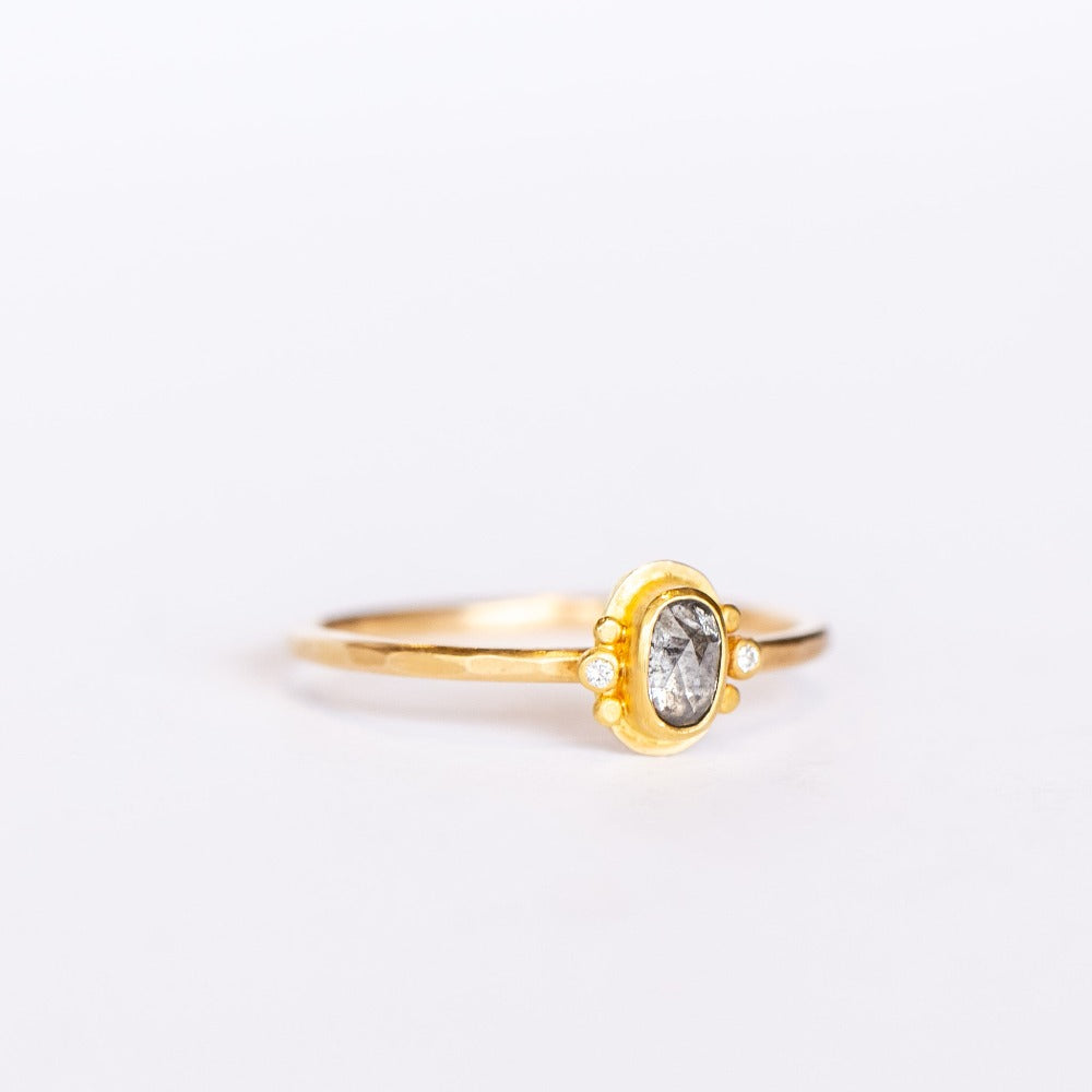 A tiny oval rose cut salt and pepper diamond is bezel set, flanked by two tiny white diamonds and dot details on a thin gold band.