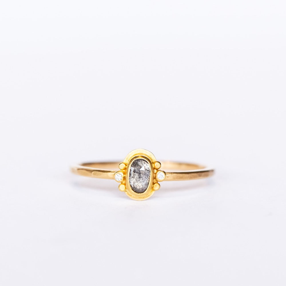 A tiny oval rose cut salt and pepper diamond is bezel set, flanked by two tiny white diamonds and dot details on a thin gold band.