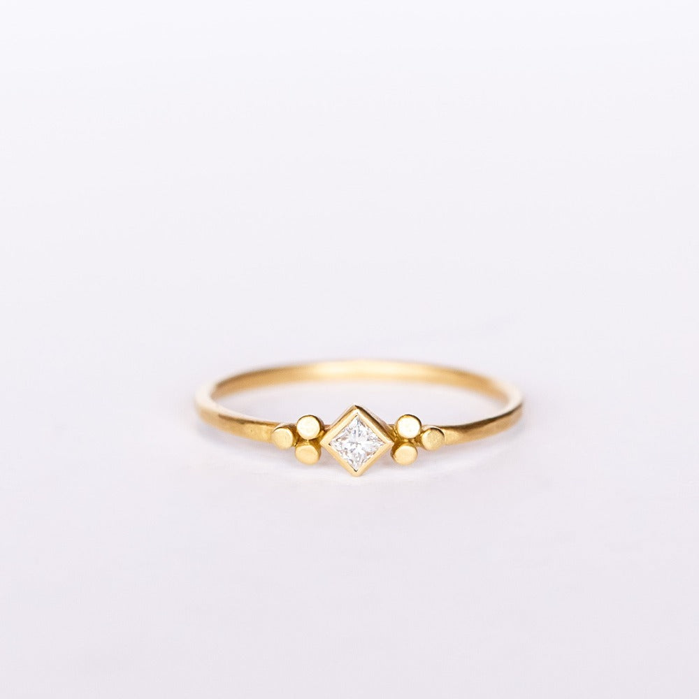 A thin gold ring featuring a kite-set princess cut diamond bezel set in yellow gold flanked by tiny dot trios.