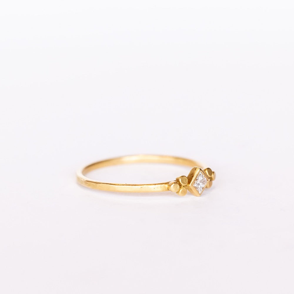 A thin gold ring featuring a kite-set princess cut diamond bezel set in yellow gold flanked by tiny dot trios.