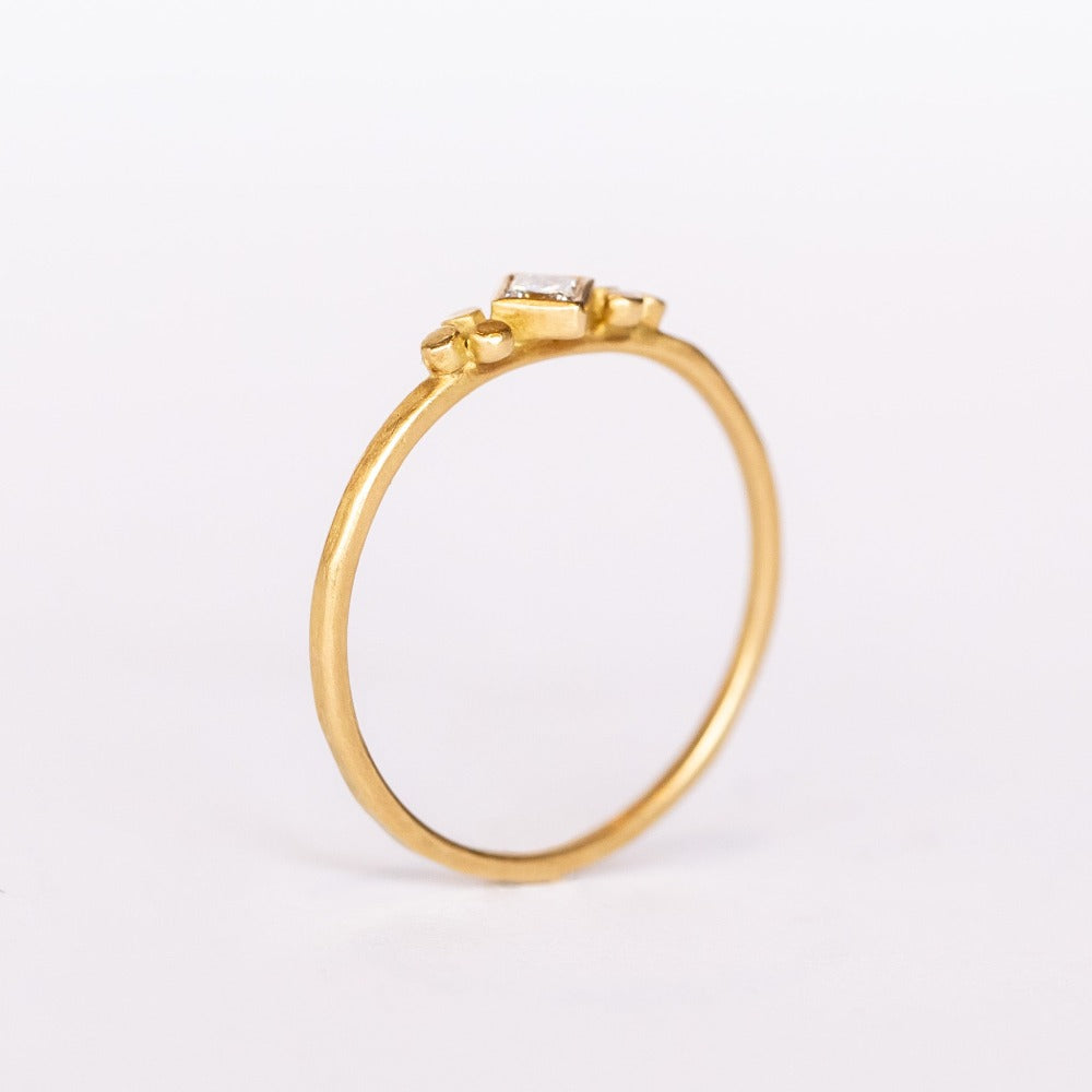 A thin gold ring featuring a kite-set princess cut diamond bezel set in yellow gold flanked by tiny dot trios.