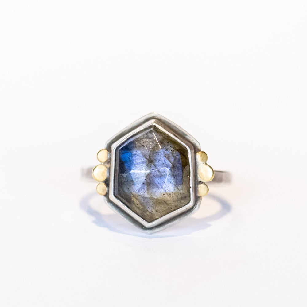 A hexagon shaped labradorite is bezel set in a sterling silver ring with tiny gold dot accents on either side.