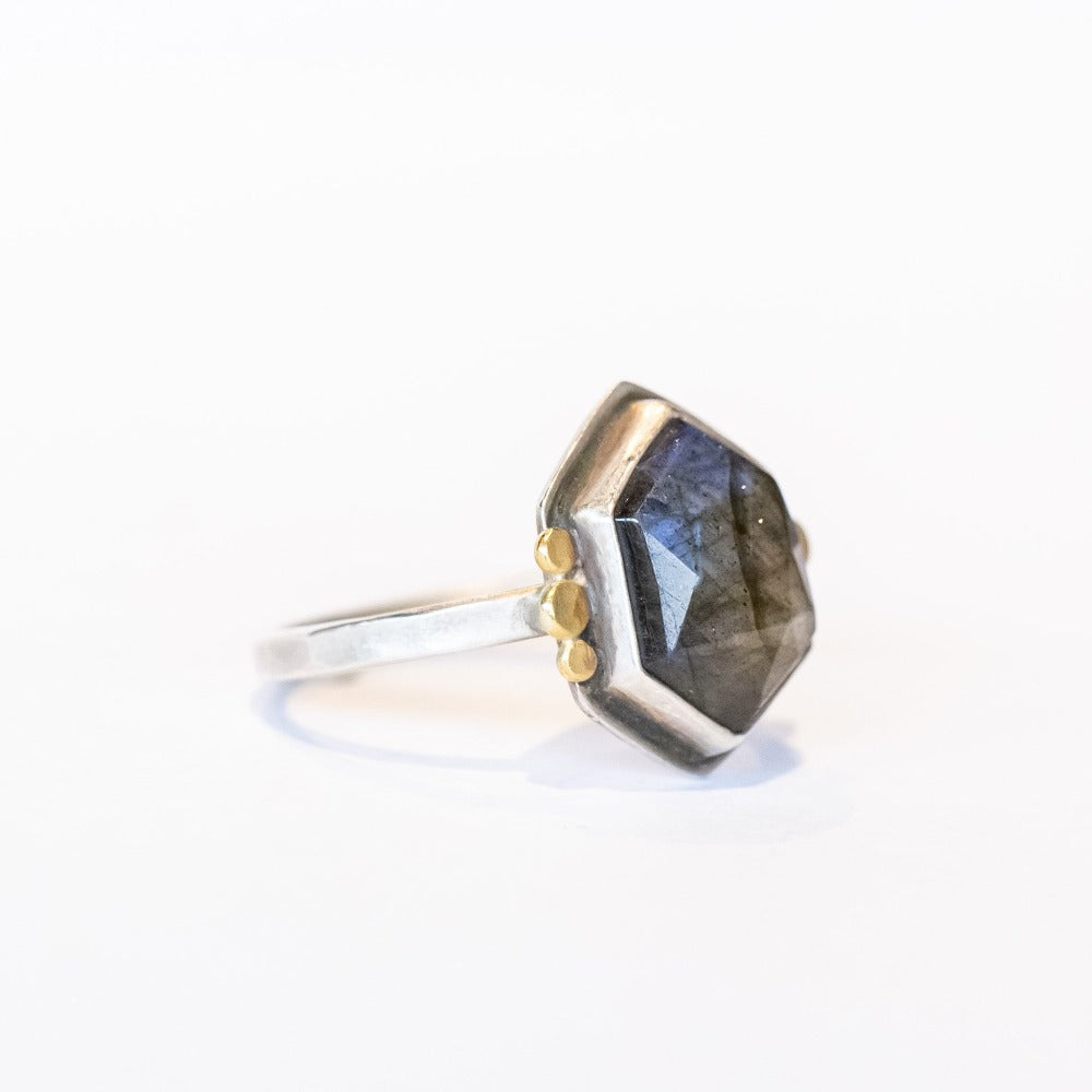 A hexagon shaped labradorite is bezel set in a sterling silver ring with tiny gold dot accents on either side.