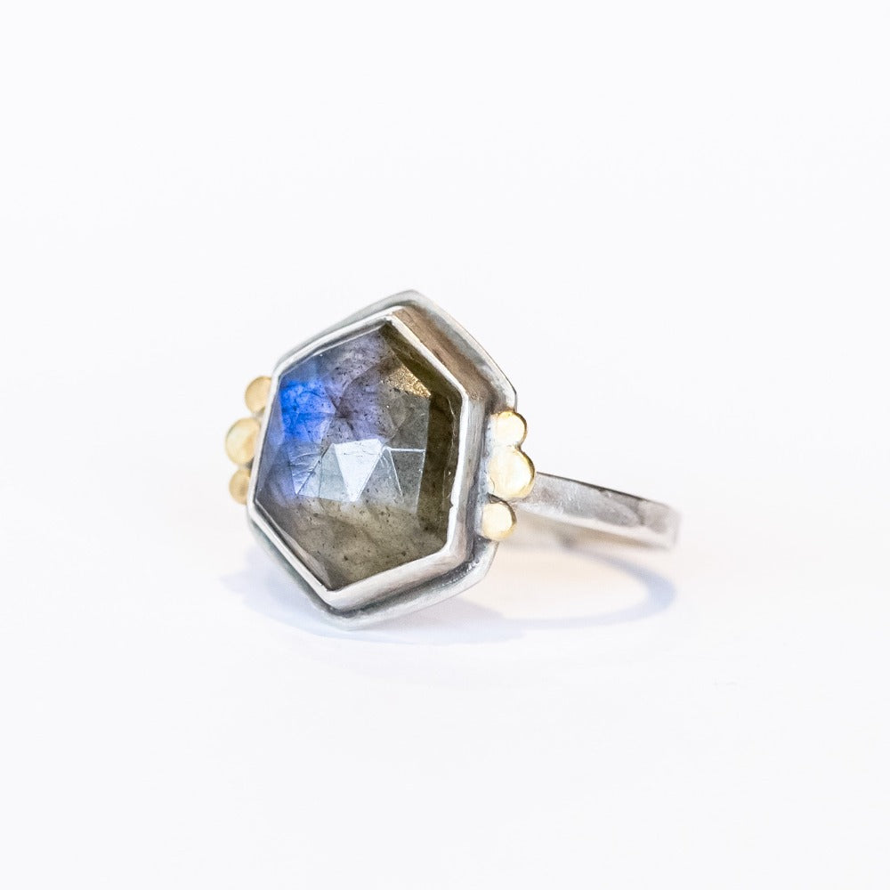 A hexagon shaped labradorite is bezel set in a sterling silver ring with tiny gold dot accents on either side.