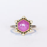 A rose cut, hexagonal shaped pink star sapphire is bezel set in yellow gold, surrounded by a halo of white diamond and yellow gold dot accents on a blackened silver ring. Front view.