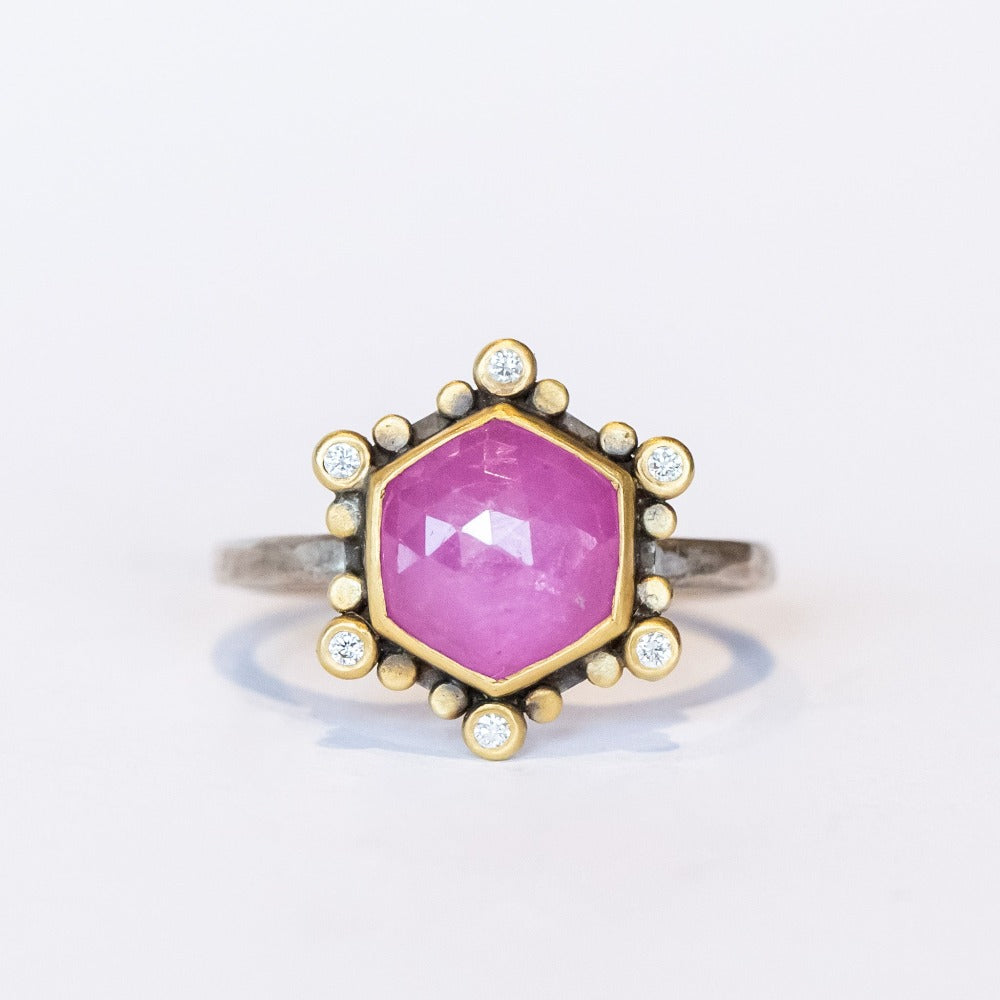 A rose cut, hexagonal shaped pink star sapphire is bezel set in yellow gold, surrounded by a halo of white diamond and yellow gold dot accents on a blackened silver ring. Front view.