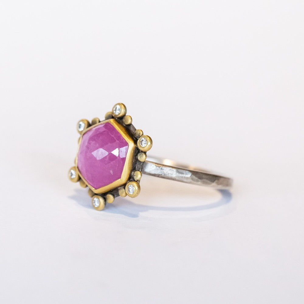 A rose cut, hexagonal shaped pink star sapphire is bezel set in yellow gold, surrounded by a halo of white diamond and yellow gold dot accents on a blackened silver ring. Side angle view.