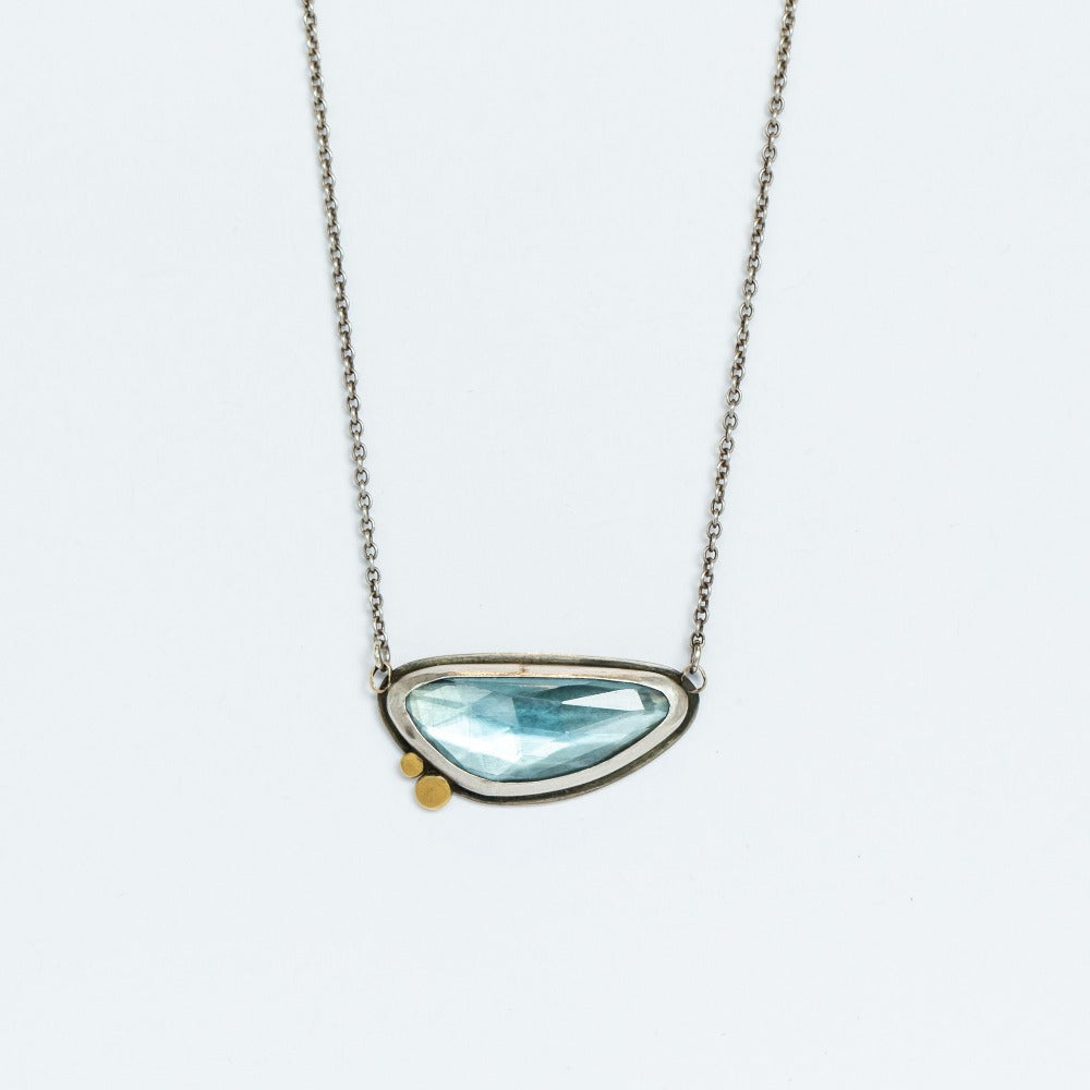 An asymmetrical, horizontal faceted blue topaz pendant set in sterling silver with two tiny gold dot accents.