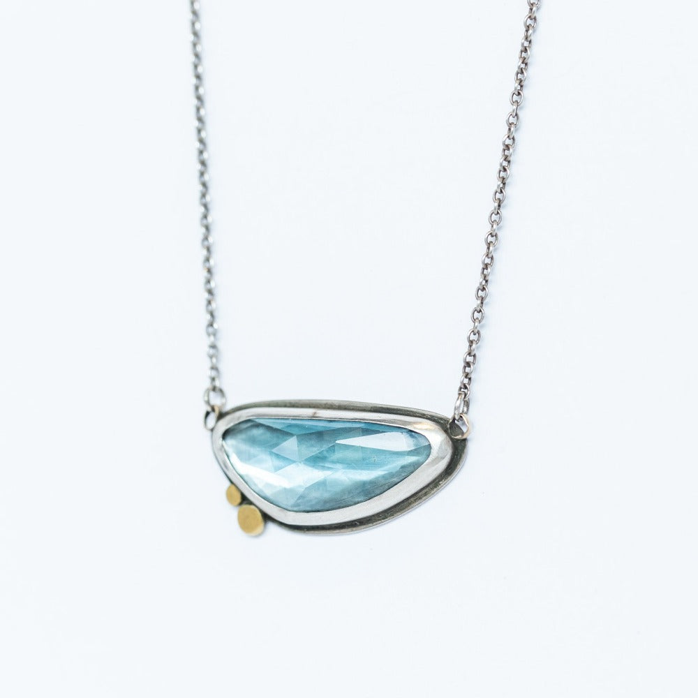 An asymmetrical, horizontal faceted blue topaz pendant set in sterling silver with two tiny gold dot accents.