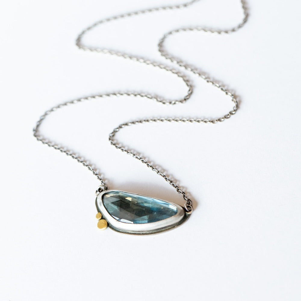 An asymmetrical, horizontal faceted blue topaz pendant set in sterling silver with two tiny gold dot accents.