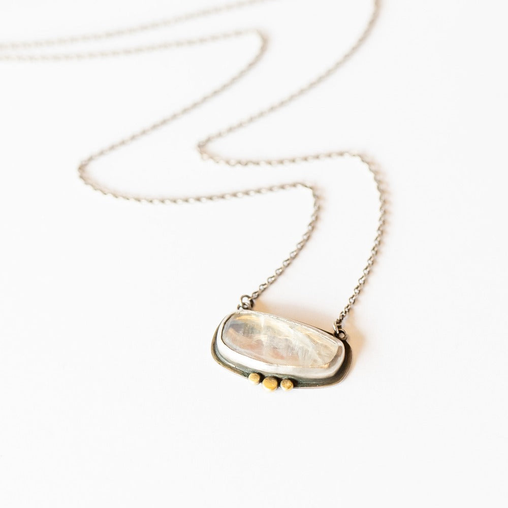 A horizontal rose cut moonstone is set in a sterling silver bezel with dainty gold dot accents along the bottom edge. It's stationed in a sterling silver cable chain necklace.