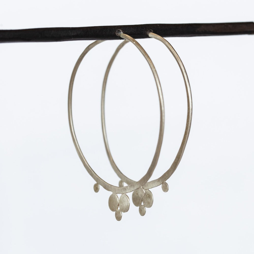 A pair of large, hammered silver hoop earrings featuring flat dot accents on the bottom edge.