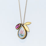 A hand painted, teardrop shape charm with a lotus is suspended from a simple cable chain with a bezel set ruby charm and petal-shaped charm.