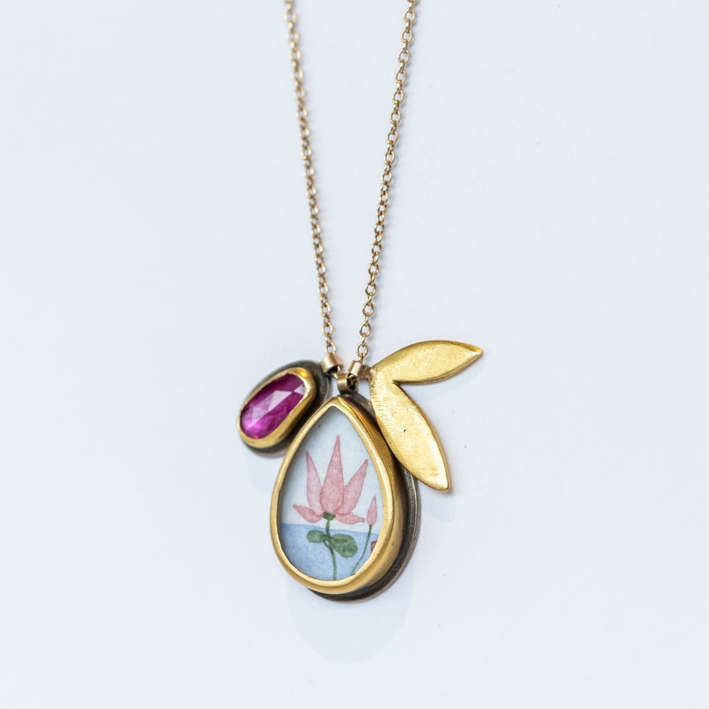 A hand painted, teardrop shape charm with a lotus is suspended from a simple cable chain with a bezel set ruby charm and petal-shaped charm.