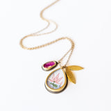 A hand painted, teardrop shape charm with a lotus is suspended from a simple cable chain with a bezel set ruby charm and petal-shaped charm.