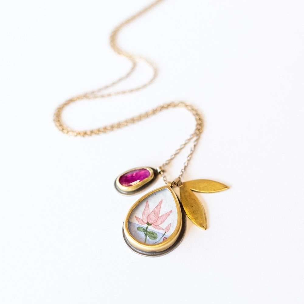 A hand painted, teardrop shape charm with a lotus is suspended from a simple cable chain with a bezel set ruby charm and petal-shaped charm.