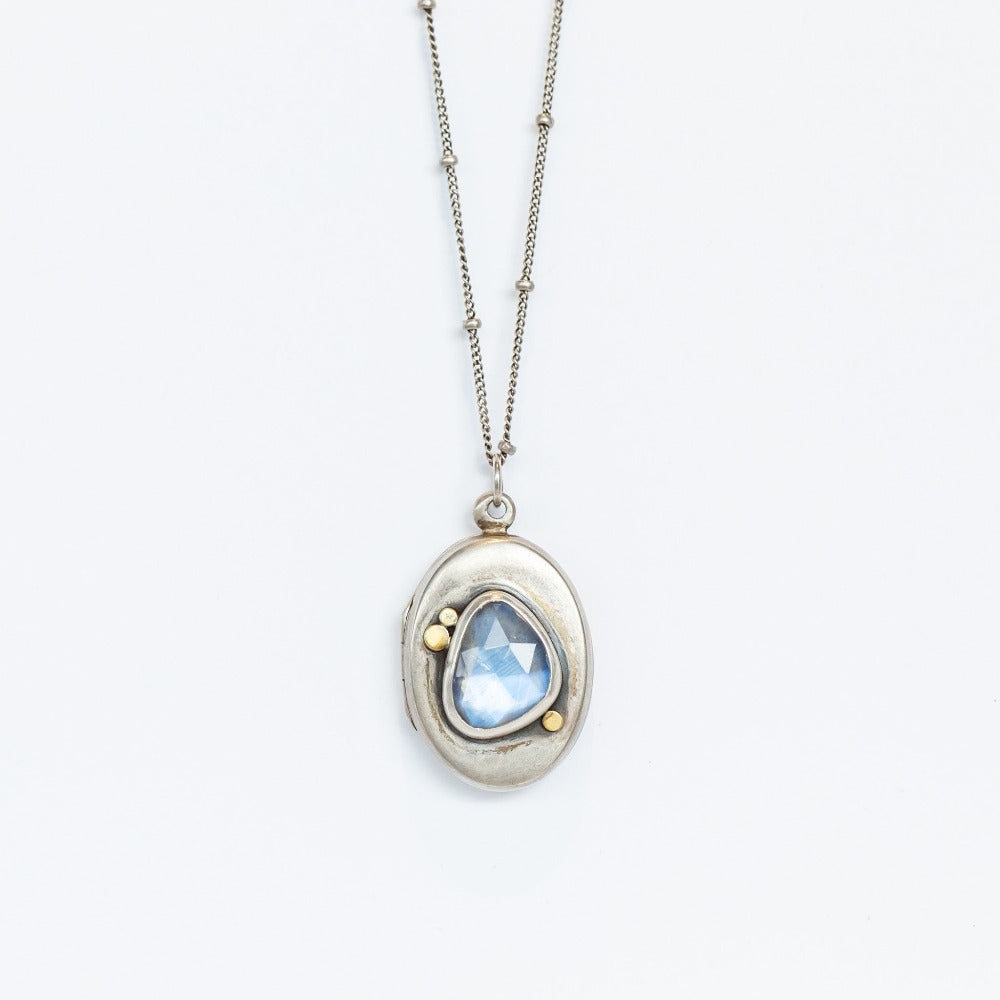 A handmade, oval shaped sterling silver locket from designer Ananda Khalsa featureing a subtly faceted asymmetrical moonstone at its center, set in sterling silver with petite 22 karat yellow gold dot details.