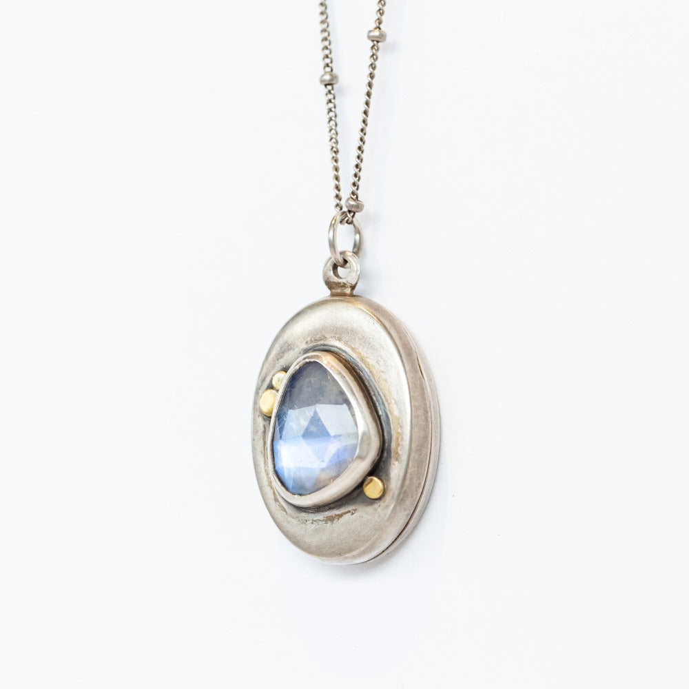 A handmade, oval shaped sterling silver locket from designer Ananda Khalsa featureing a subtly faceted asymmetrical moonstone at its center, set in sterling silver with petite 22 karat yellow gold dot details.