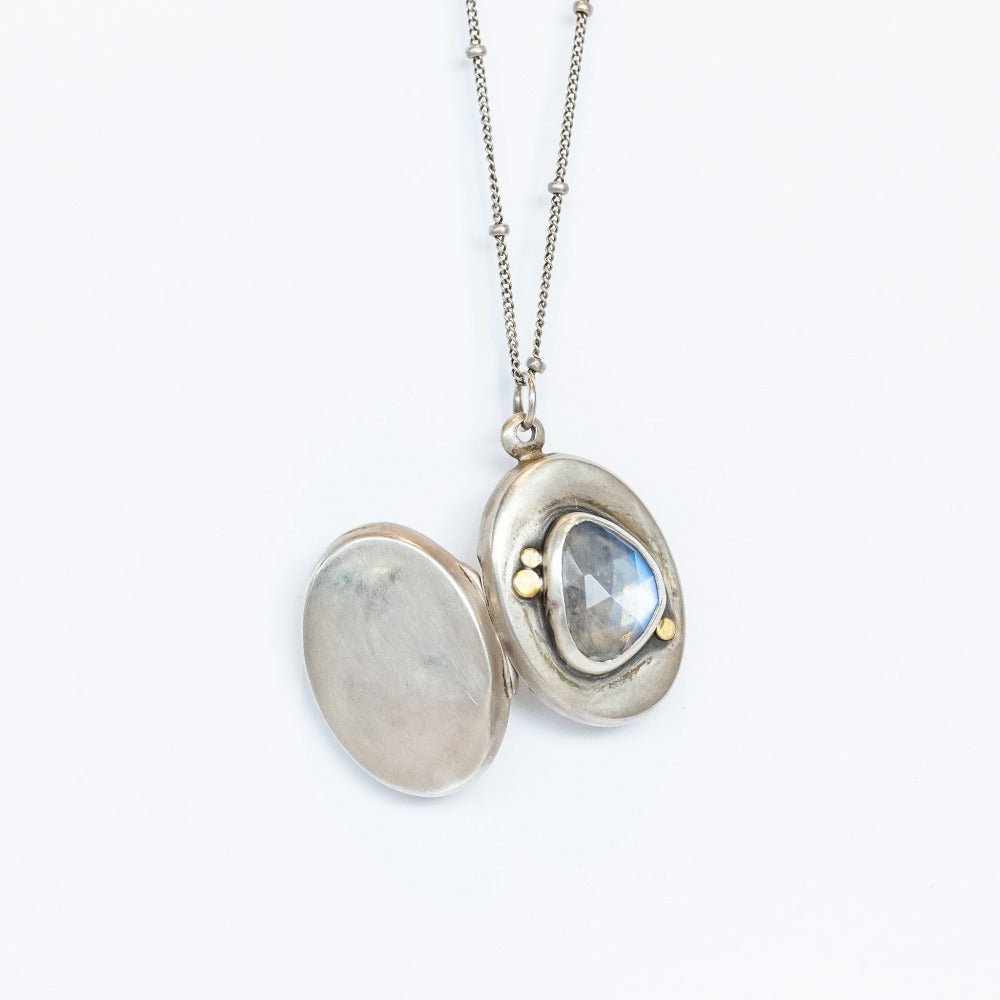 A handmade, oval shaped sterling silver locket from designer Ananda Khalsa featureing a subtly faceted asymmetrical moonstone at its center, set in sterling silver with petite 22 karat yellow gold dot details.