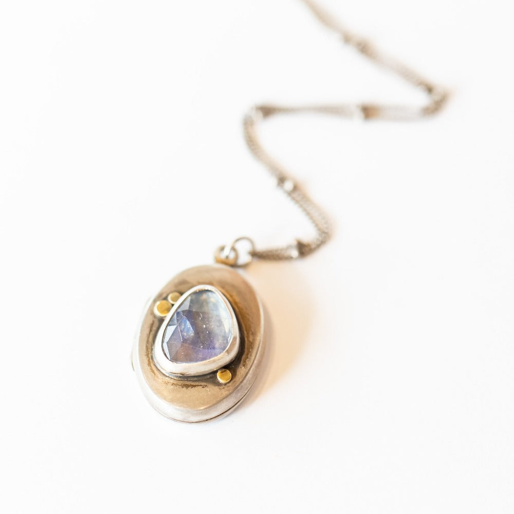 A handmade, oval shaped sterling silver locket from designer Ananda Khalsa featureing a subtly faceted asymmetrical moonstone at its center, set in sterling silver with petite 22 karat yellow gold dot details.