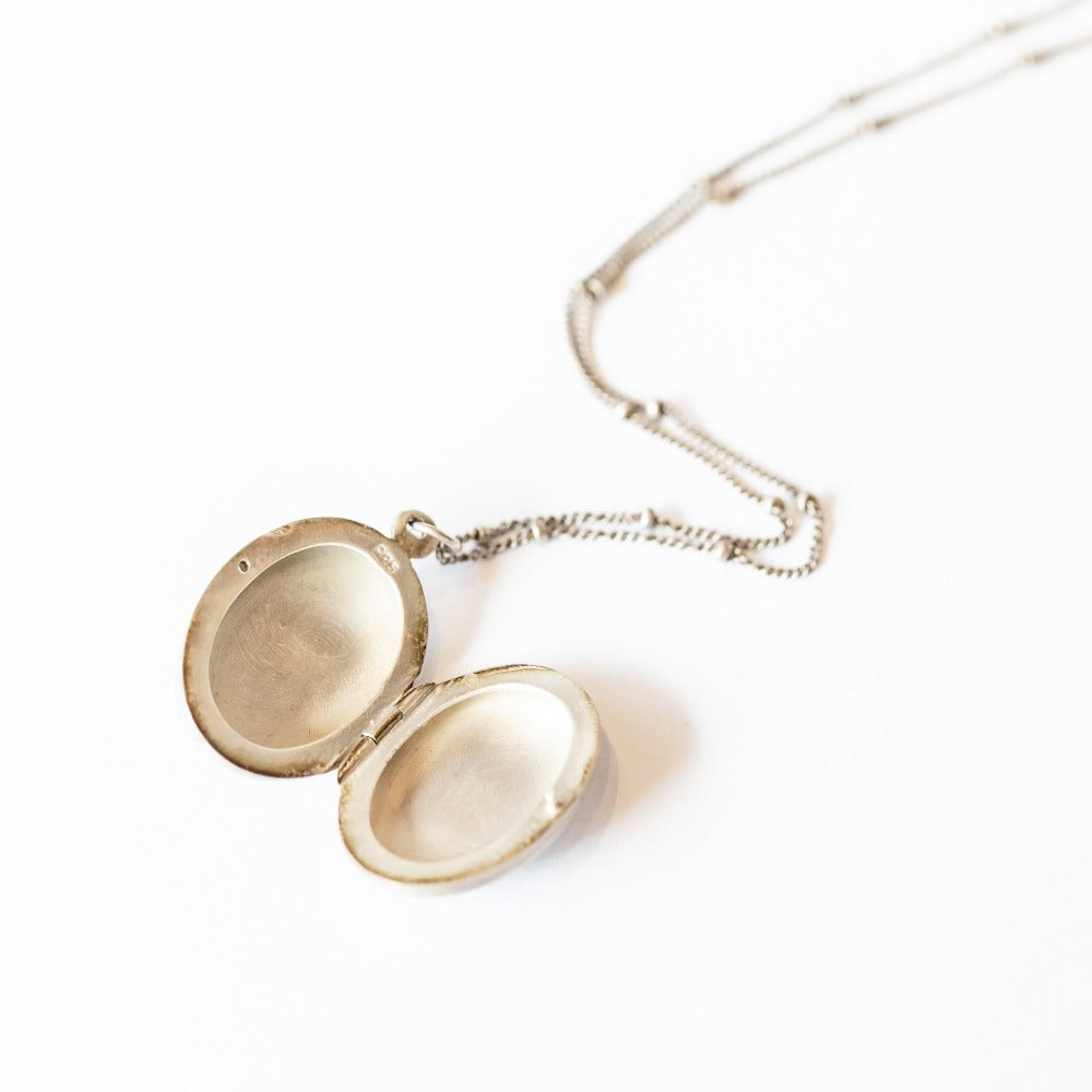 A handmade, oval shaped sterling silver locket from designer Ananda Khalsa featureing a subtly faceted asymmetrical moonstone at its center, set in sterling silver with petite 22 karat yellow gold dot details.