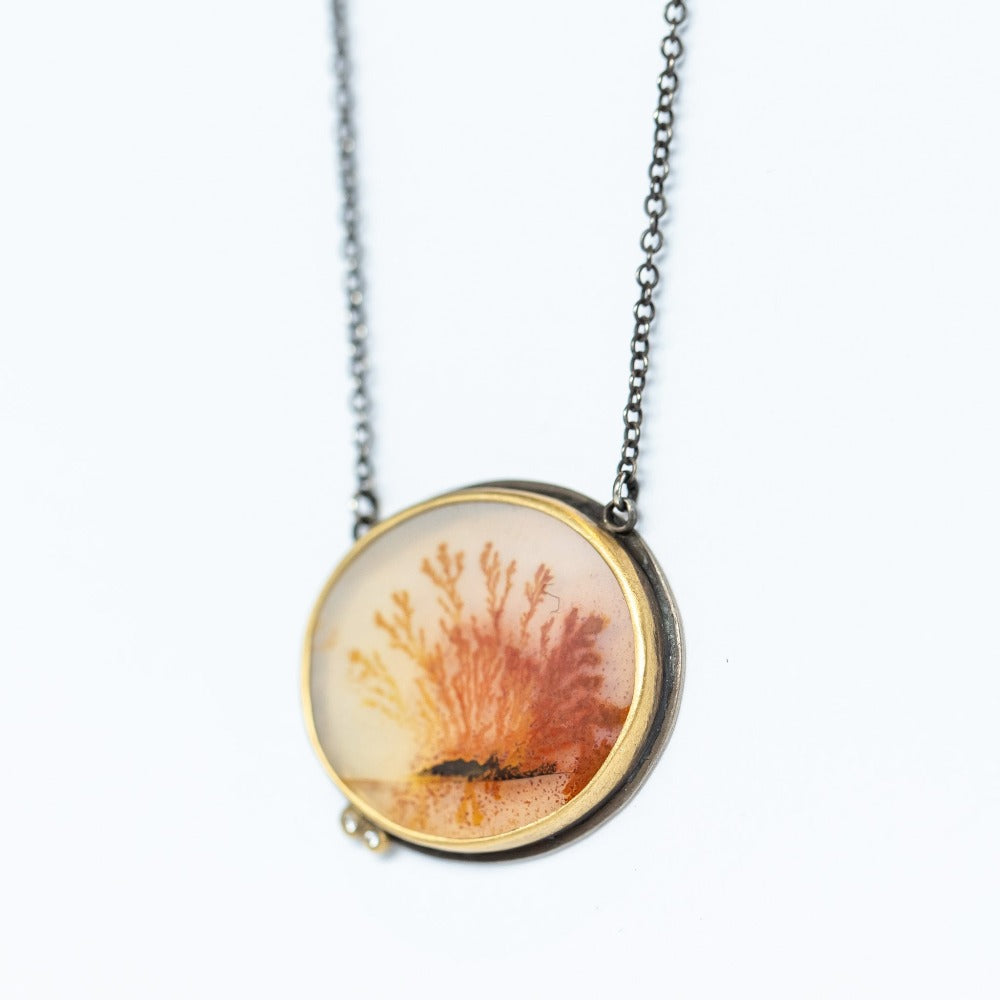 An oval shaped, orange dendritic agate is bezel set in yellow gold with two diamond accents. Necklace is sterling silver.